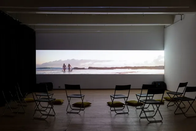 Video install playing in a space with empty chairs and cushions.