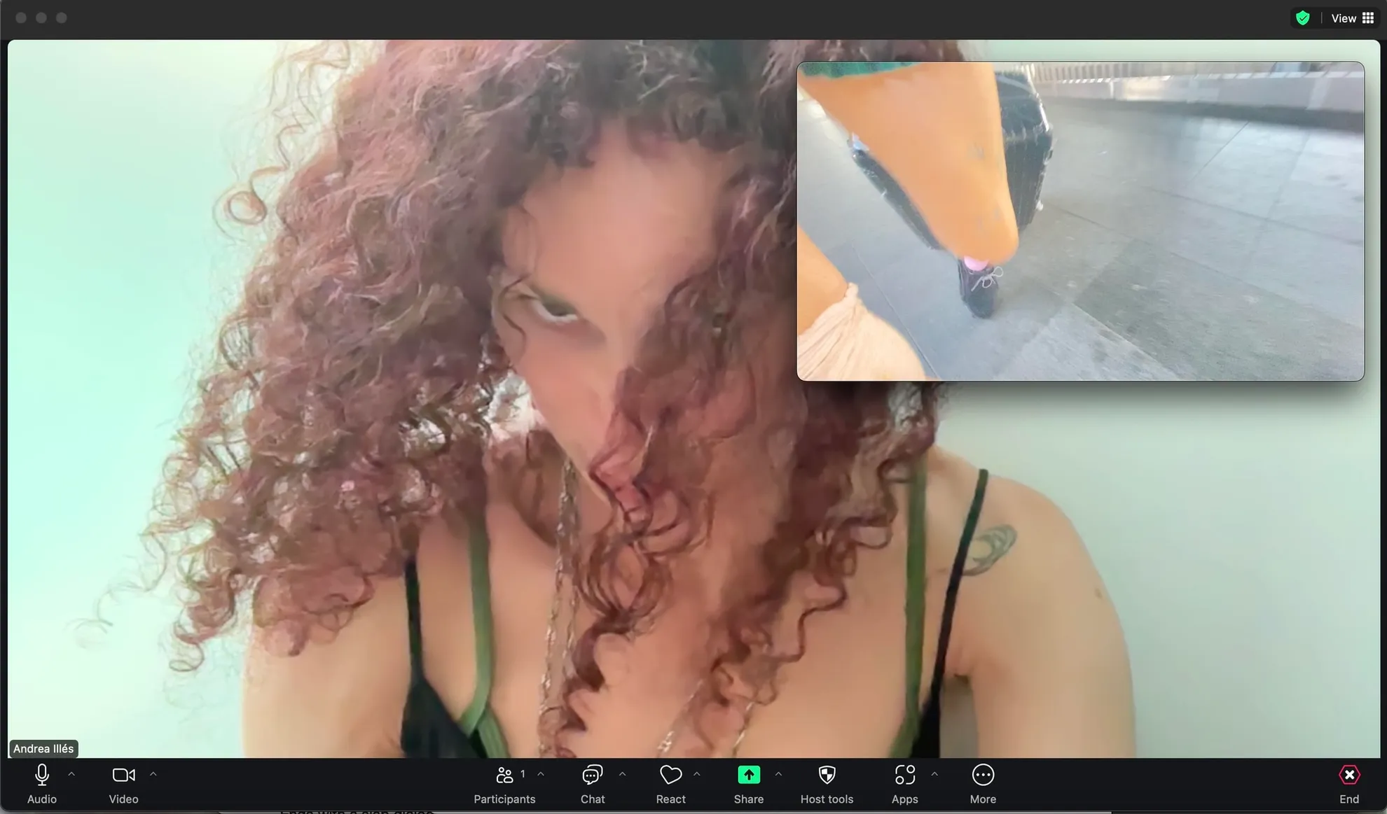 A screen shot of a woman on a zoom call. She has a strappy singlet and long dark red curly hair. There is a window within the window of a womans leg while she walks.