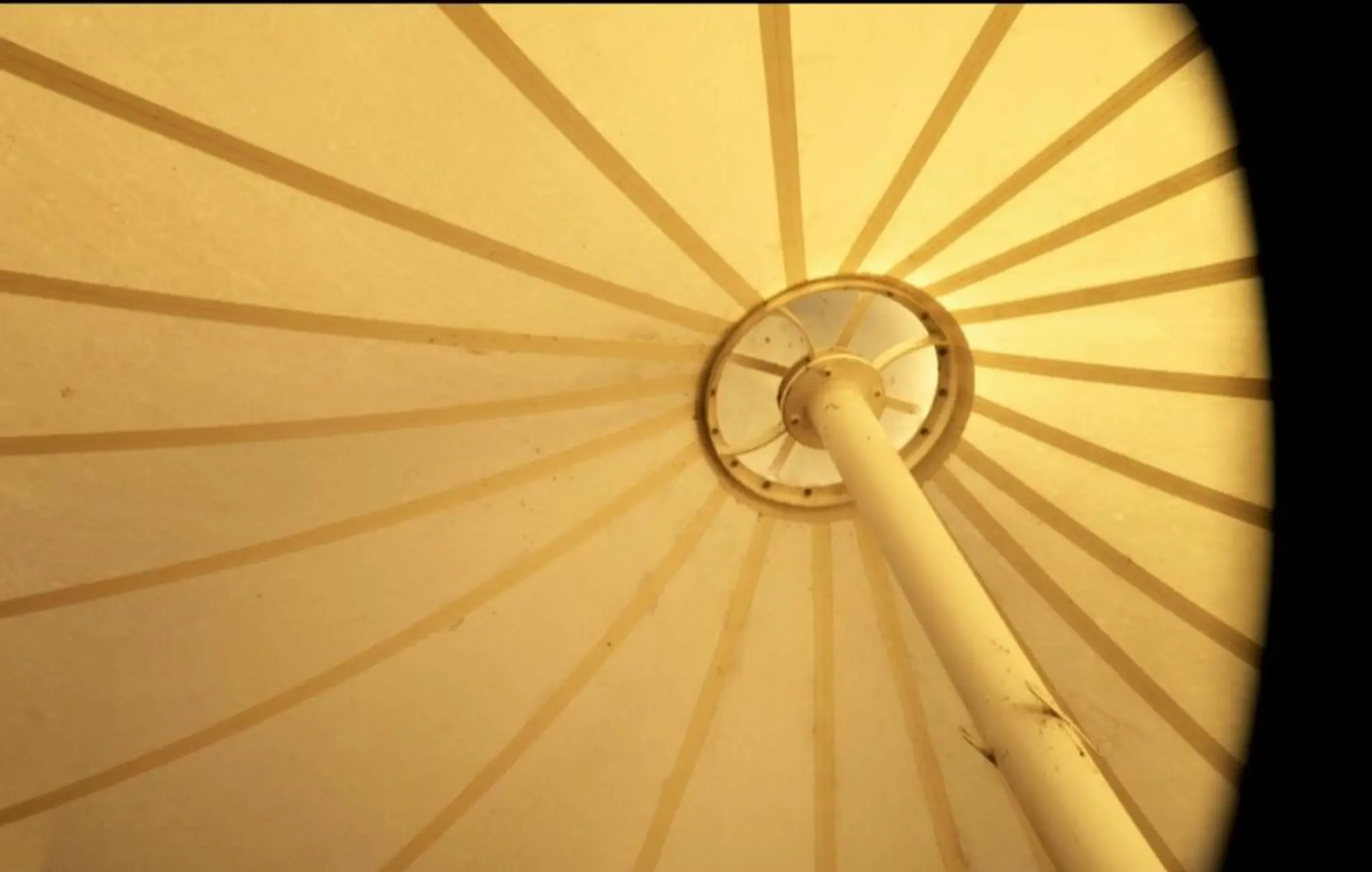A film still of the inside roof of a tent, stretched taught and held up by a white metal pole. The light is tinted yellow, and the fabric joins in the tent create a stripy pattern that fans out from the centre point.