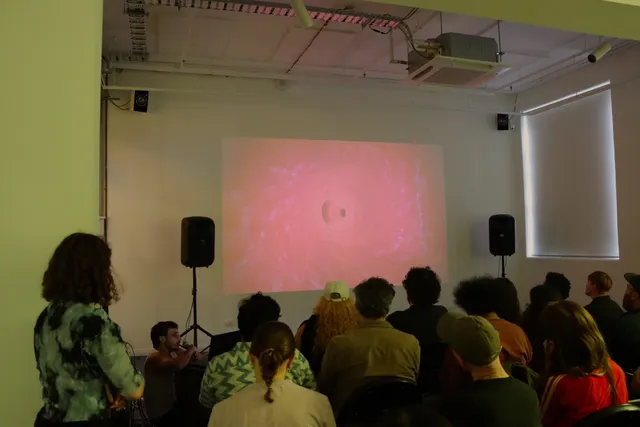 A group of people sitting on chairs watching Joel Sherwood Spring's videowork projected onto a white wall.