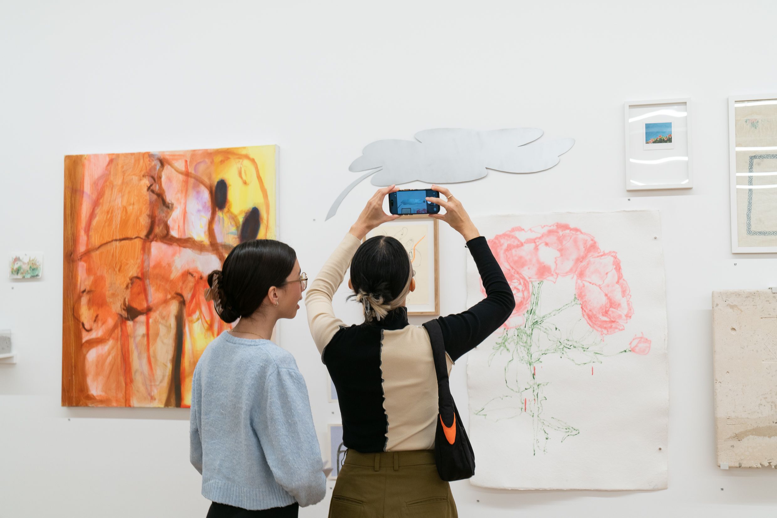 Two figures standing in front of a collection of artworks hanging on the wall. The figure on the left is holding up their phone to take photos.
