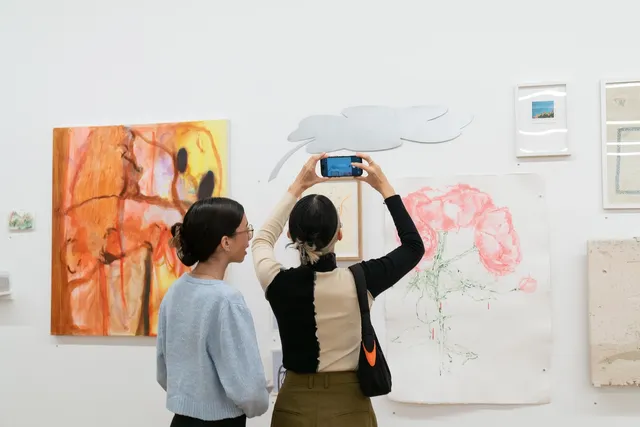 Two figures standing in front of a collection of artworks hanging on the wall. The figure on the left is holding up their phone to take photos.