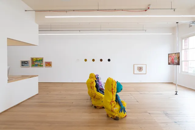 A wide angle installation view of the 'Slime and Ashes' exhibition at West Space. Terry Williams' colourful fabric sculptures are sitting on the floor in the front and centre and Victoria Todorov's video wok on the right with. Lionel Grijvala's three paintings, Tony Albert's five glass cast faces and Bronwyn Hack's framed underwear against the white wall in the background.