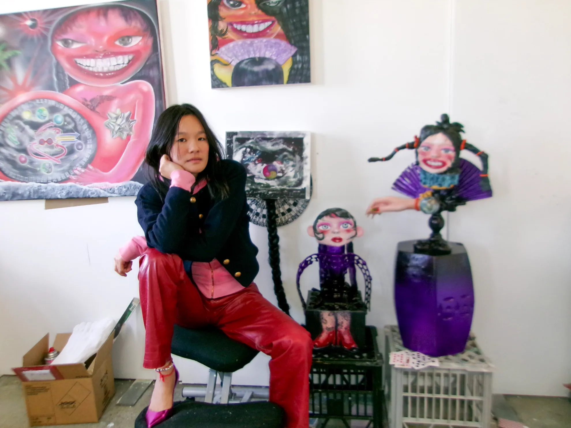 The artist Jacquie Meng sitting on a chair posing for a photo amongst a collection of their paintings and sculptures.