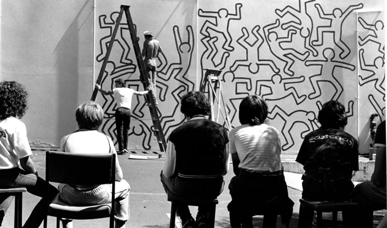 The back of some people sitting on the chairs looking at two people with ladder in front of a wall painted with human shapes.