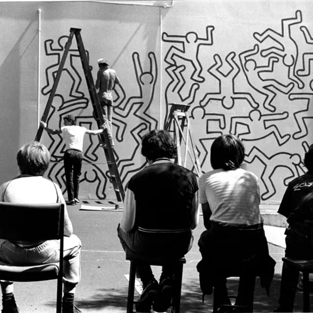 The back of some people sitting on the chairs looking at two people with ladder in front of a wall painted with human shapes.
