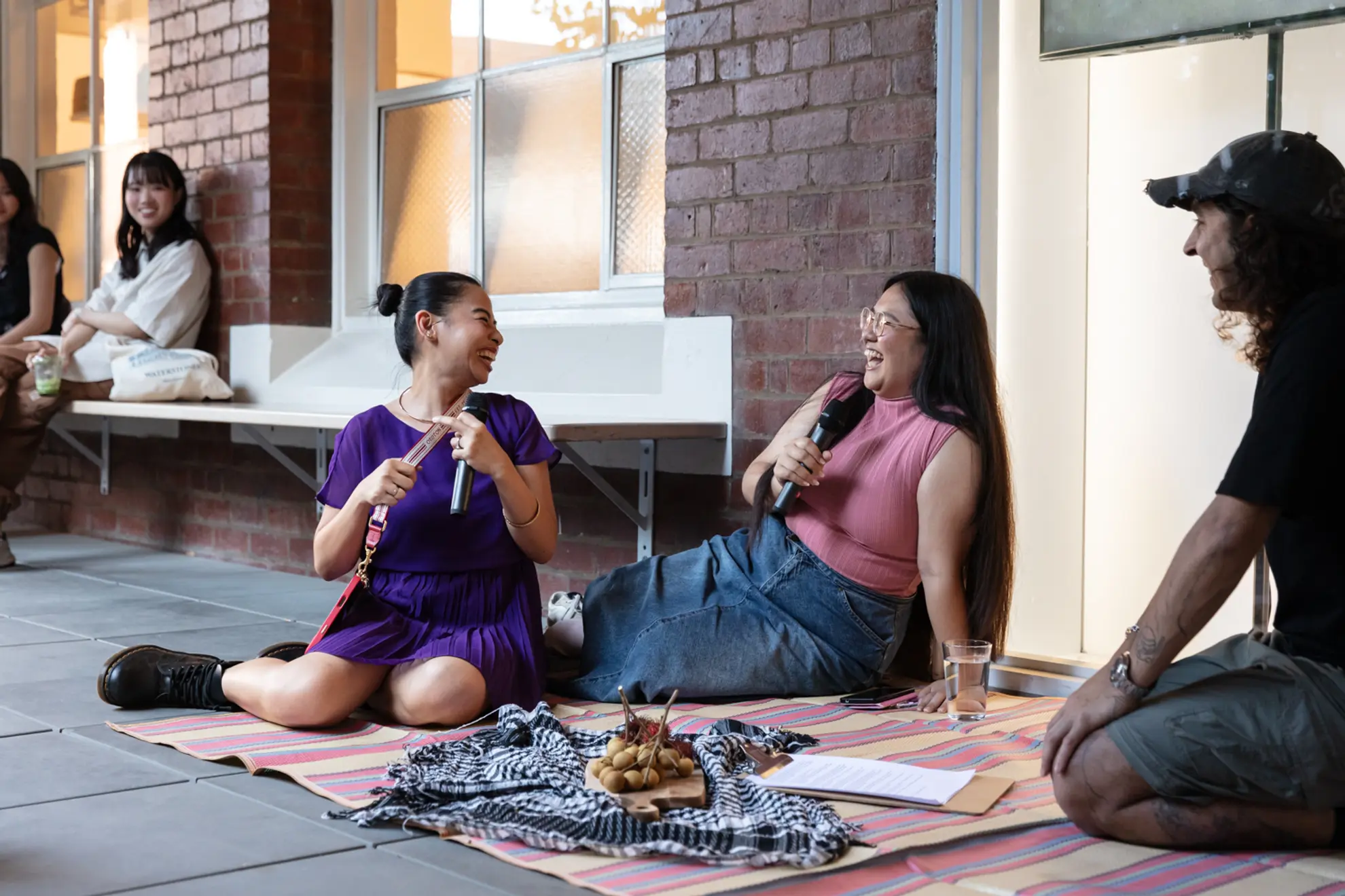 An individual in a purple dress sits with their legs folded, beside two other figures on a rug. One in black and the other in a pink shirt. The one in pink is holding a microphone smiling back at the person in purple, whose holding a microphone and some kind of lenyard. There are hints of a crowd as two figures behind them look on smiling, on benches.The 3 main figures are on a rug situated in an open walkway lined with large white windows and some brick.