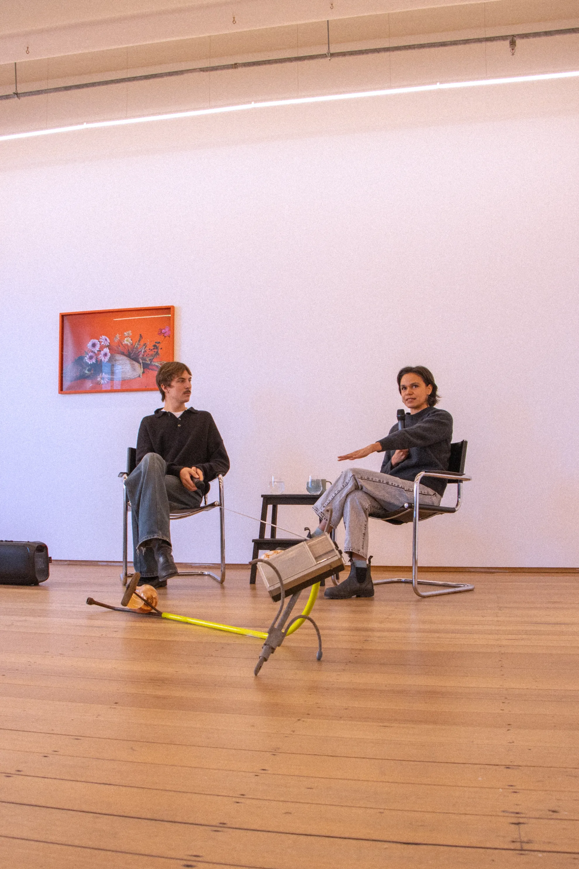 Michael and Katie are seated in conversation. Katie is mid speech holding the microphone, on the wall behind Michael is a framed photographic work of Katies of flowers resting on a shell, in the foreground lies a sculptural installation piece of metal and a radio.