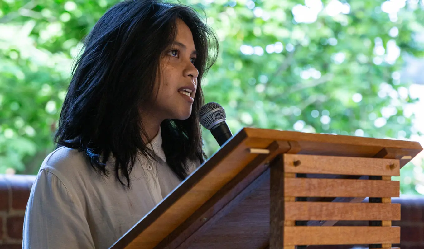 A girl holding a microphone behind a wooden platform holding text, pears upward as they talk. Behind there is a large green tree, out of focus. They are depicted from the knees up.