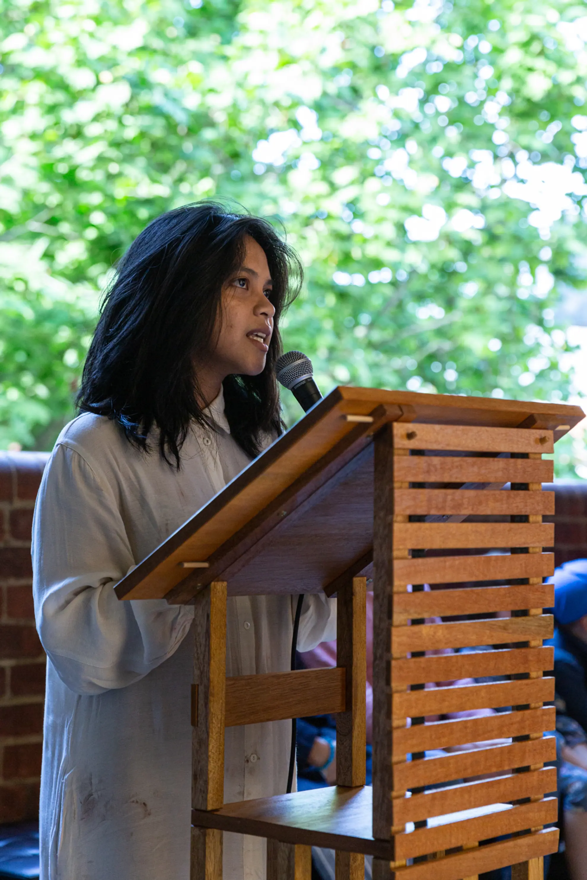 A girl holding a microphone behind a wooden platform holding text, pears upward as they talk. Behind there is a large green tree, out of focus. They are depicted from the knees up.