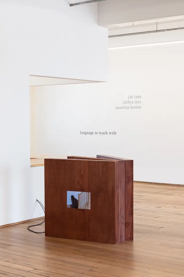 A wooden frame structure sits within the gallery space on the floor with a small screen playing a video work. The background features the west space gallery wall with the exhibition title language to reach with and the artist names of chi tran, nithya Iyer, and moorina bonini.