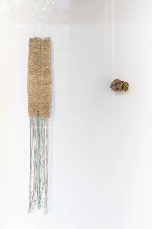 A long weaving with long weft threads hanging off the bottom and a lump of unfired wild clay infused with plastic gems hang on the wall of the West Space Window gallery