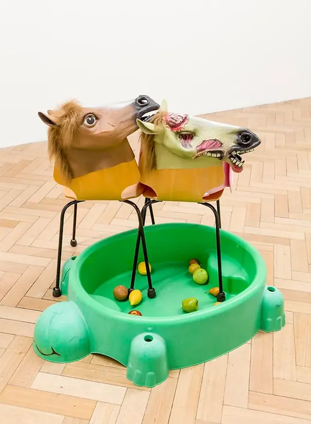 3 horse masks rest on two wooden chair tops, like bodies to the horse. 3 our of 8 of the chair legs dip their 'feet' into a green turtle pool that is holding 10-15 fruits and apples