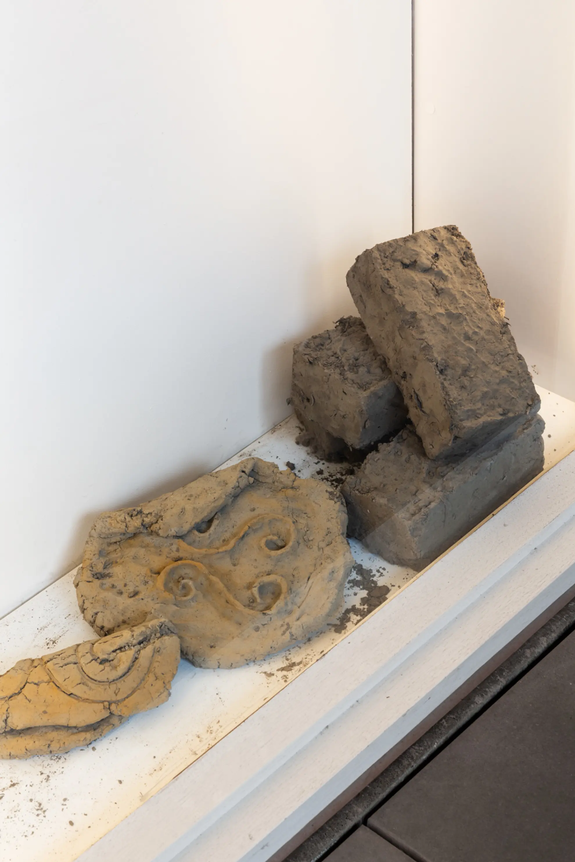 An installation view of several clay works and some hand made mud bricks sit in the base of the West Space Window gallery. There are spirals engraved and embossed in some of the clay works