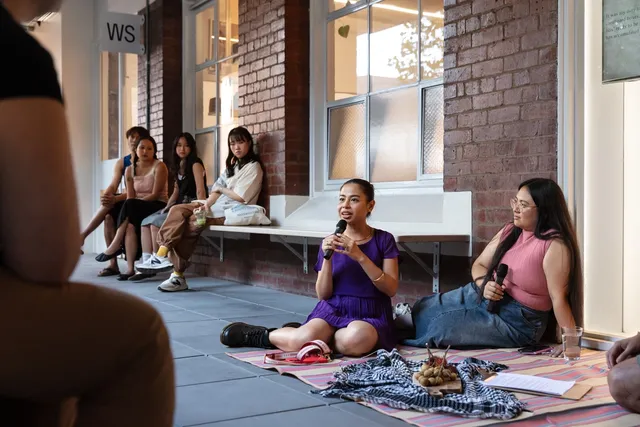 A group of people line the edges of an open corridor listening to some figures speak. The main speaking figure is wearing deep purple holding a microphone as they smile and gaze around at the people listening. Another figure with long brown hair in pink stares, listening, to the talking person. Theyre legs are folded. In front of them is a cloth holding some fruit-like forms on a chopping board, assumably informing their discussion.