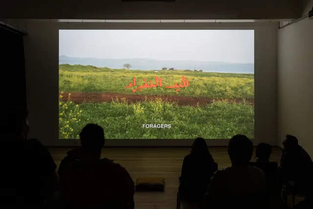 People sitting in a dark room watching a film projected on a white wall. The red Arabic word appears on the background of grass landscape. The white subtitles writes "foragers".