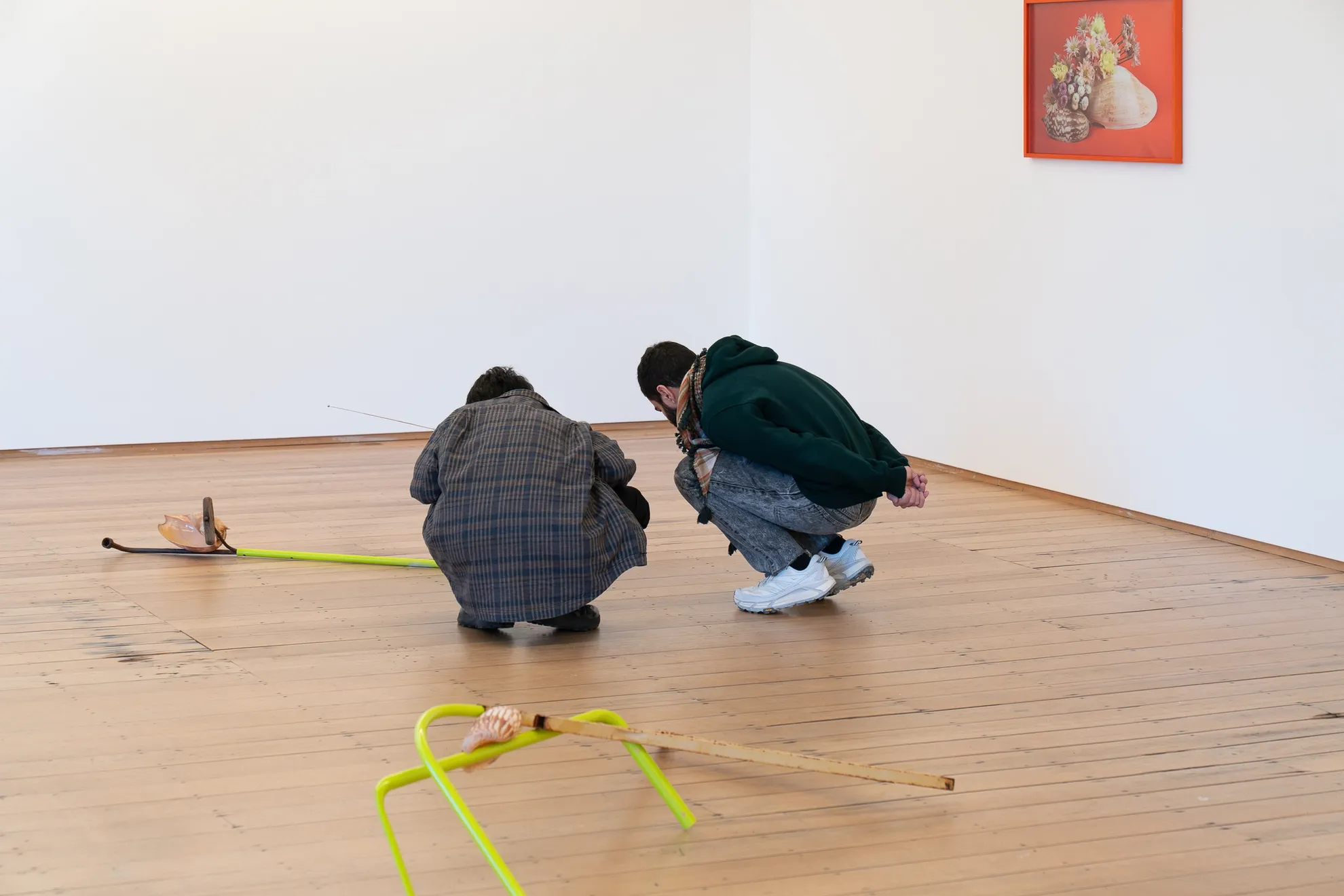 Two people squat close to one of the sculptural installation pieces on the floor of the gallery, we see metal poles and shells, a second installation is in the foreground. On the wall behind them hangs a framed photographic piece of flowers resting on a shell.
