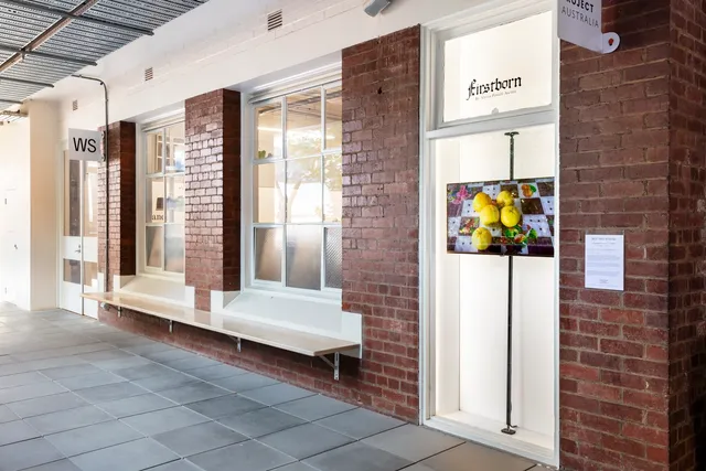 An install shot of the corridor out the front of West Space. There are two large windows lined with white skirting and the narrow window exhibition platform. In the window platform, a screen displays an image of 5 yellow fruits on a checkered table cloth. The screen is installed on a thick vertical beam for structural integrity.