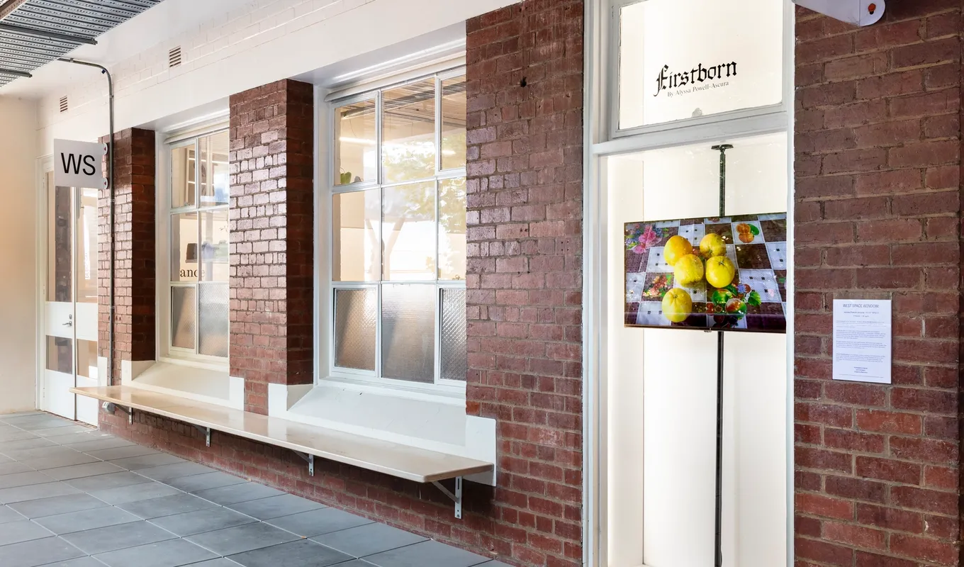 An install shot of the corridor out the front of West Space. There are two large windows lined with white skirting and the narrow window exhibition platform. In the window platform, a screen displays an image of 5 yellow fruits on a checkered table cloth. The screen is installed on a thick vertical beam for structural integrity.