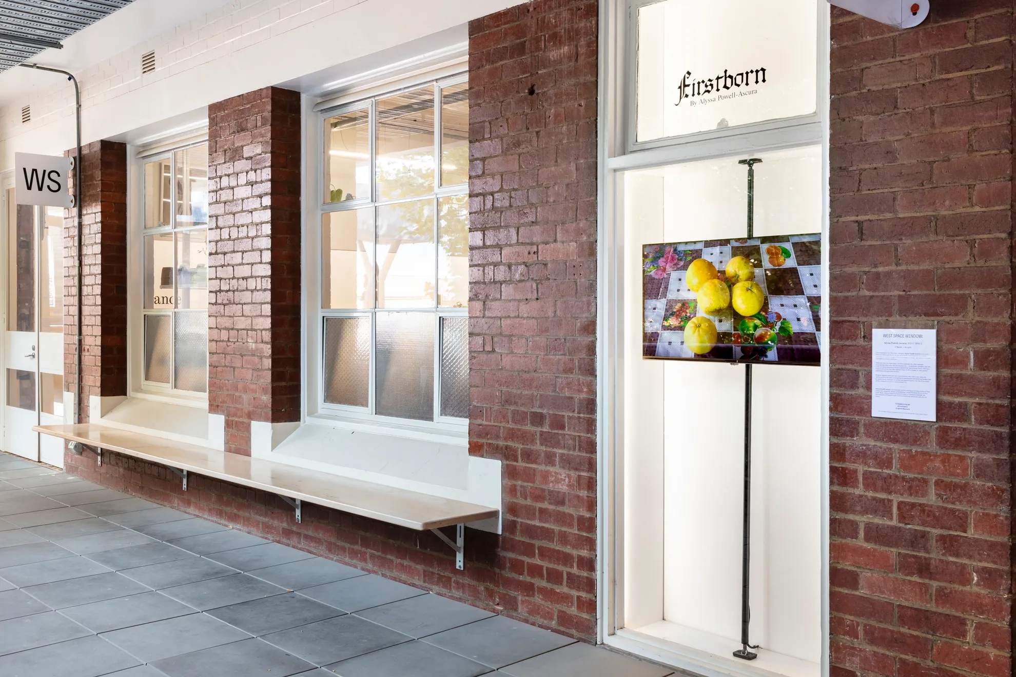An install shot of the corridor out the front of West Space. There are two large windows lined with white skirting and the narrow window exhibition platform. In the window platform, a screen displays an image of 5 yellow fruits on a checkered table cloth. The screen is installed on a thick vertical beam for structural integrity.