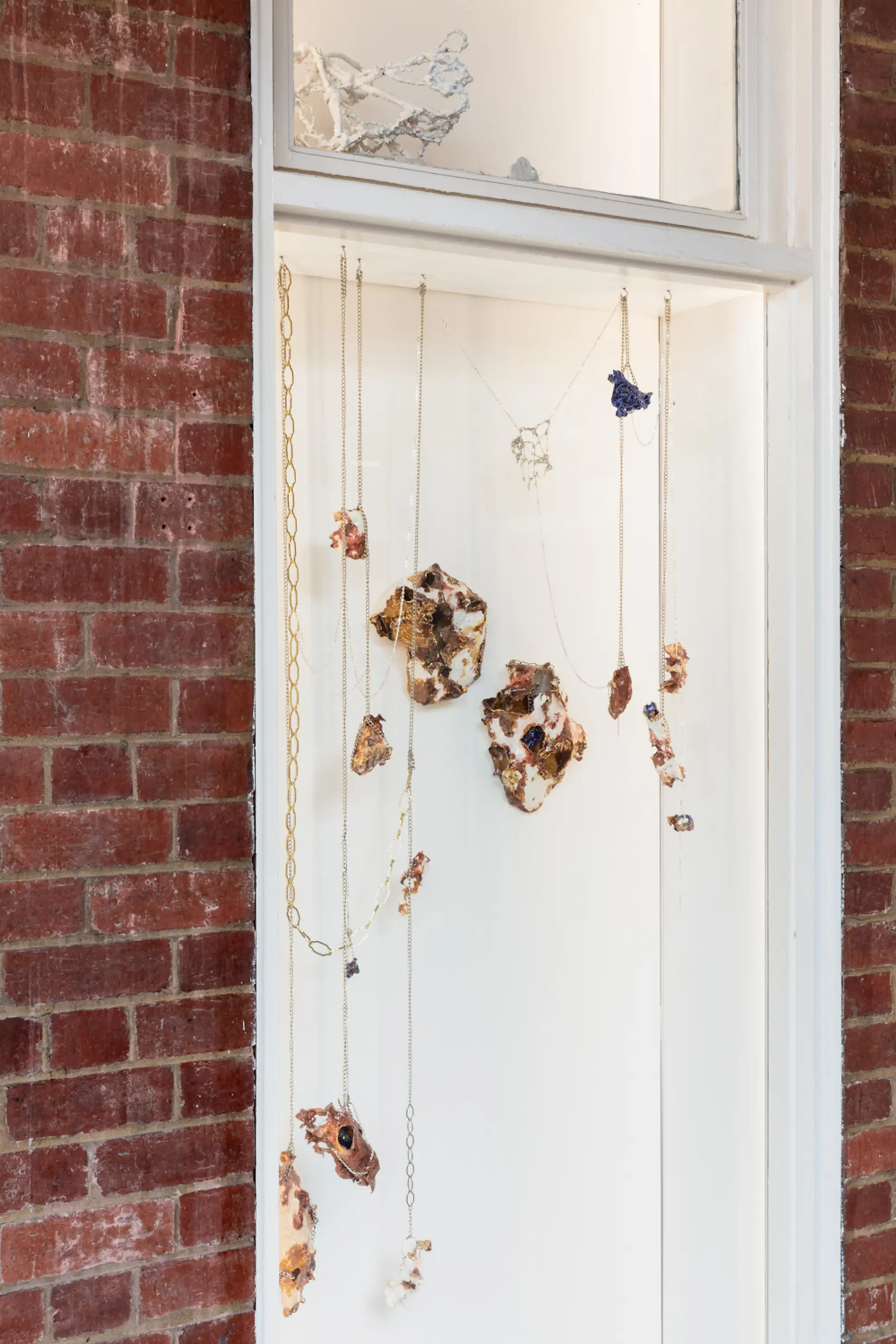 A series of marbled porcelain and ceramic sculptures hang from chains in a white window which is a recess in a red brick wall.