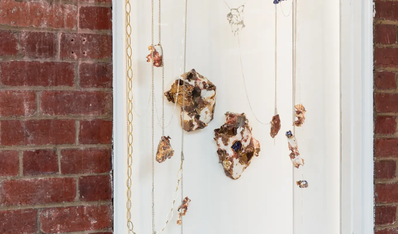 A series of marbled porcelain and ceramic sculptures hang from chains in a white window which is a recess in a red brick wall.