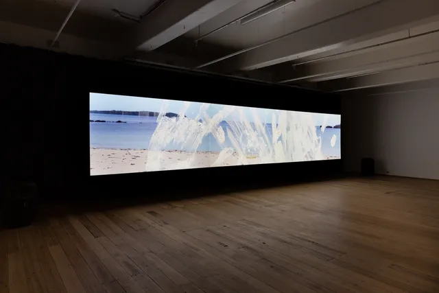 A film is projected in a long rectangular shape on the galley wall. The film still pictures a beach with hand prints in white paint layered on top of the image.