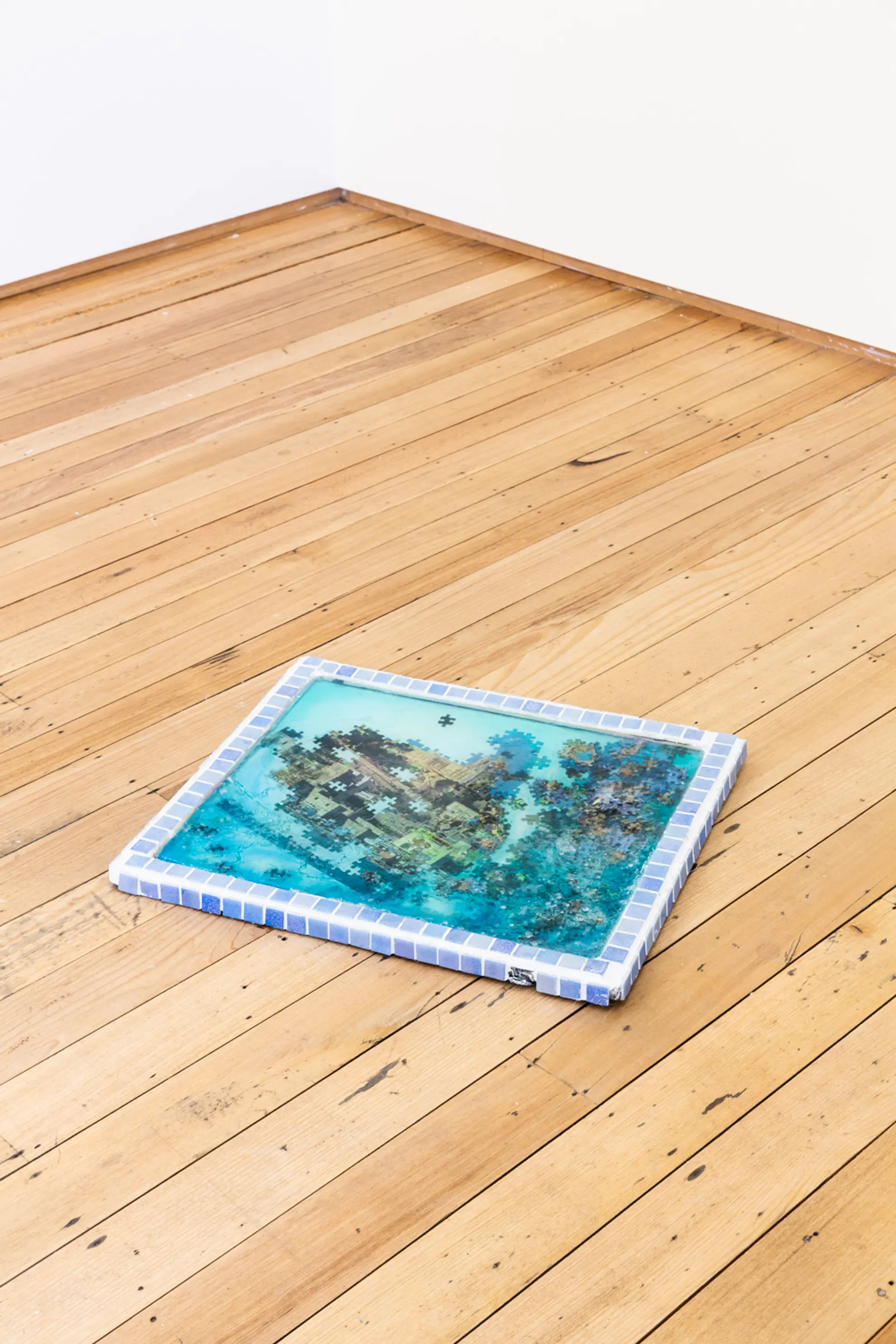 A rectangular sculptural artwork made of a border of blue tiles and filled with puzzle pieces suspended in blue resin sits in the corner of a room on the floorboards