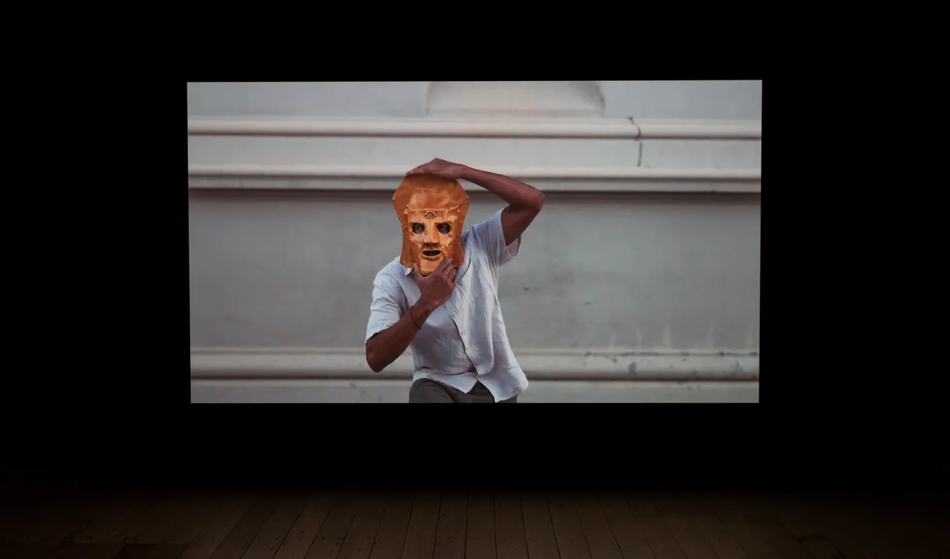 A video still from a large projection hangs in a dark room. In the video, a man wearing a white shirt holds a large decorative mask over his face.