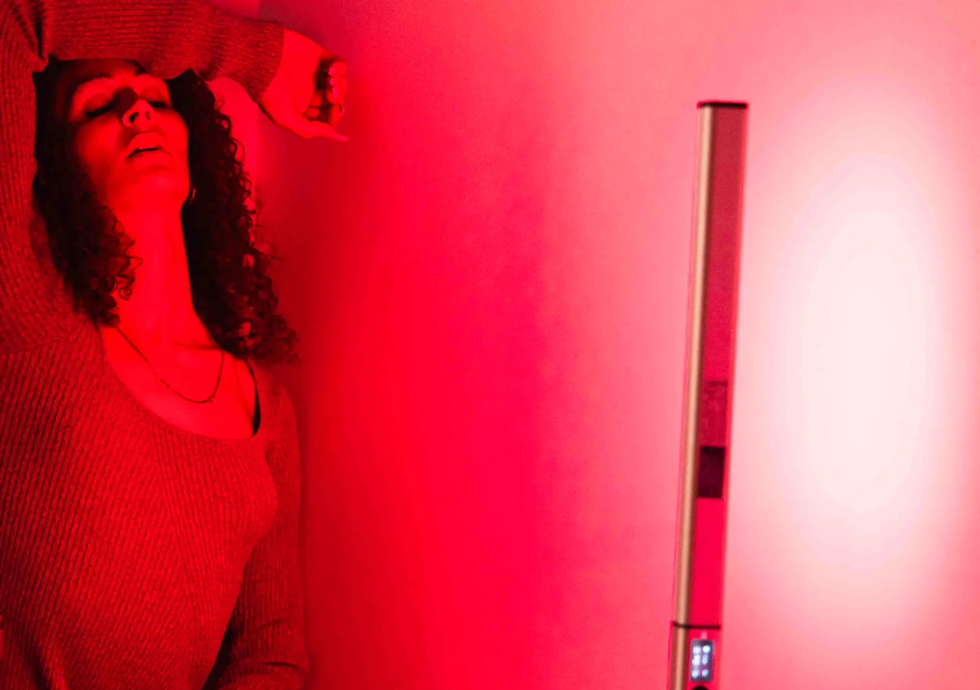 A person with dark curly hair and a knitted jumper stands in a room with a red hue, they rest their forearm across their head.