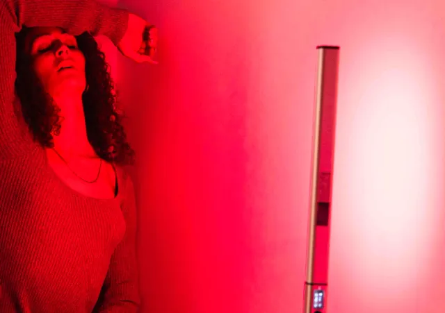 A person with dark curly hair and a knitted jumper stands in a room with a red hue, they rest their forearm across their head.