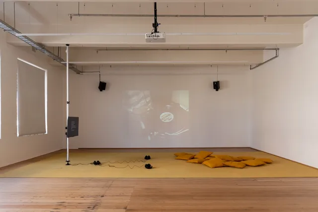 Installation View of 13 Years showing a screen suspended away from the Gallery wall with 3 pairs of headphones and some cushions in front of it. The back wall shows a projection of archival footage of a record sitting on a record player with a person standing over it.