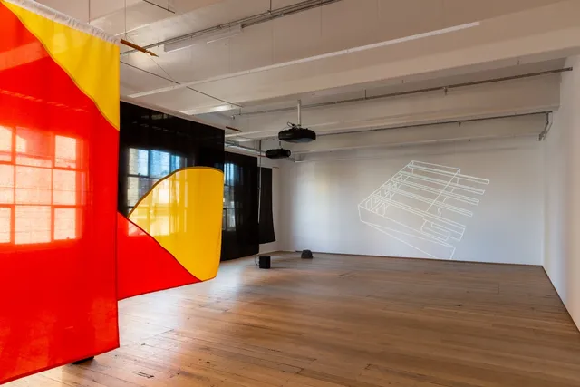 Installation view of "Big Pharmakon". The image shows 4 semi-transparent fabric sheets hanging in front of 3 windows in the West Space Gallery. The first sheet is partially visible and is red with a small triangular yellow section in the top right. The second shows a segment of a yellow circle bisected by a red triangle in the bottom left over a black background. The third and fourth sheets are completely black. On the ground in front of the sheets are two speakers. The back wall shows a projection of a representation of a large building-like structure.