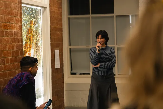 Person standing with microphone giving a speech and another person watching with brick walls and windows behind them. One large window brightly lit displaying colourful print and hanging orange flowers.