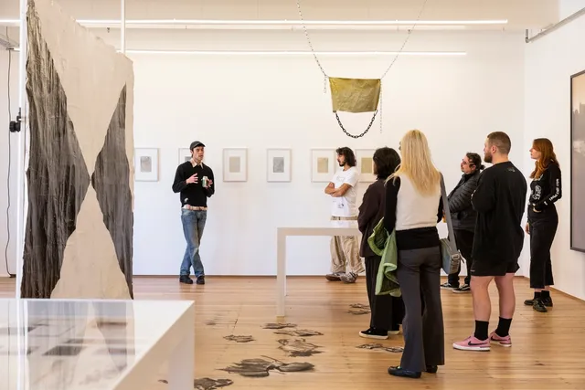 A crowd gather in an exhibition space listening intensely to an individual talk, gesturing toward the work. There around around 7-8 people present.