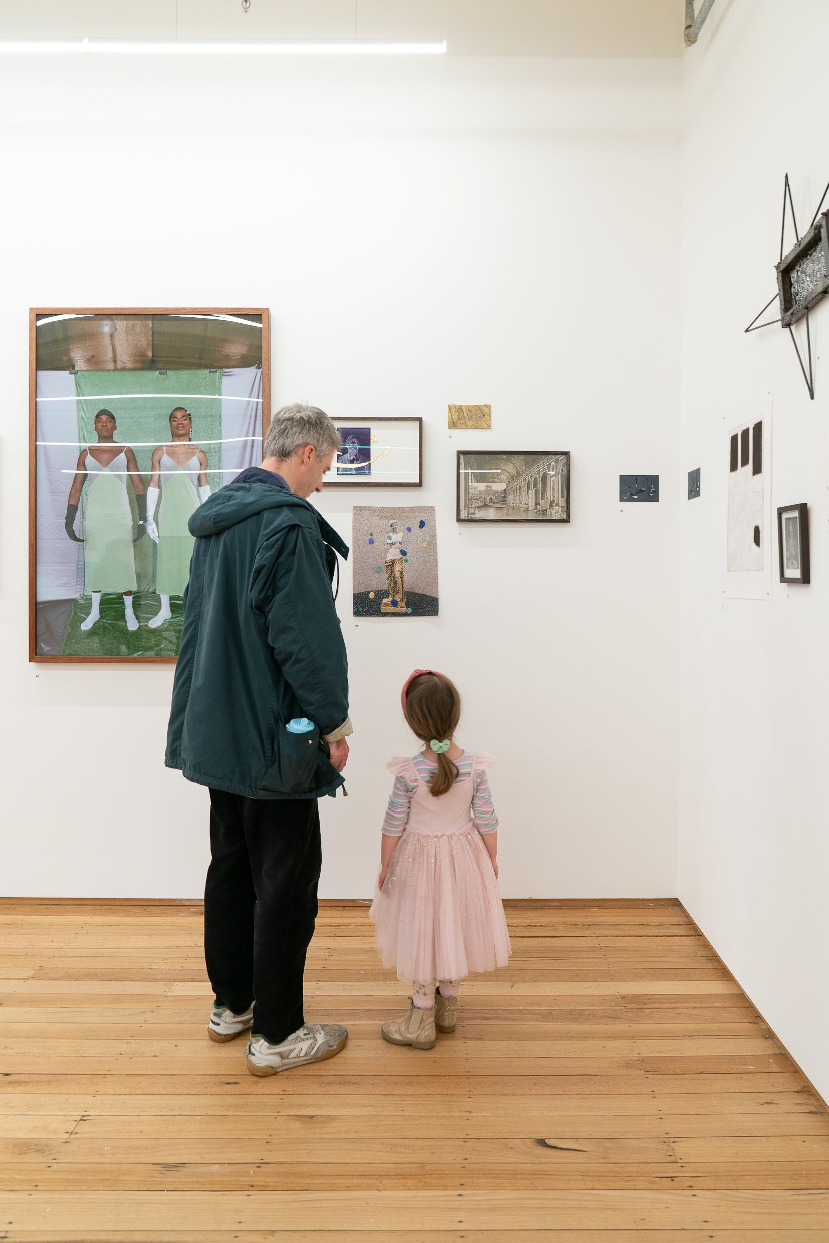 A man and a girl standing side by side viewing a collection of artworks hanging on the wall.