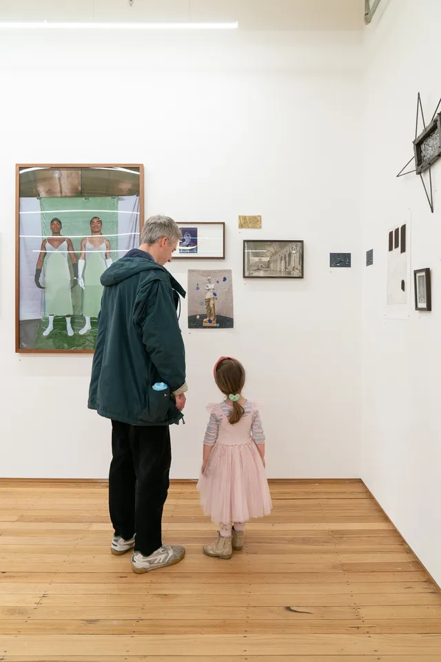 A man and a girl standing side by side viewing a collection of artworks hanging on the wall.