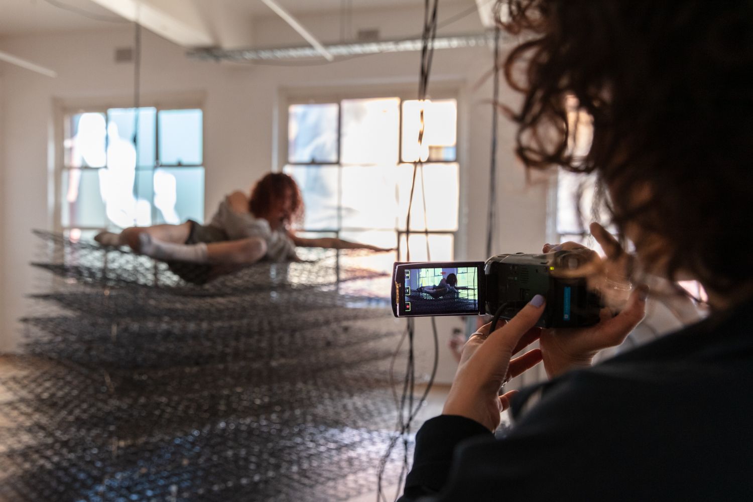 A woman is lying on a pile of metal springs of mattresses. A person holding a video camera is recording.