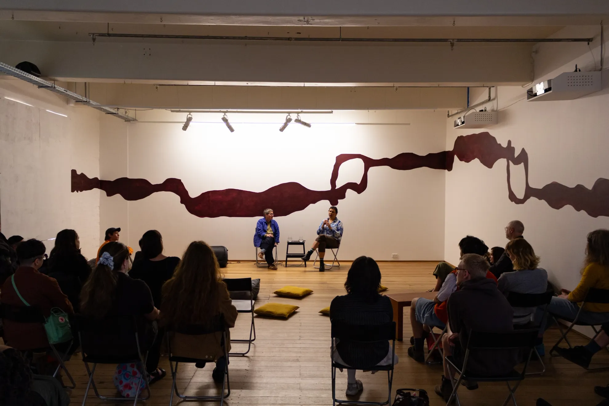 Two individuals sit in a gallery space crossed-legged on two folding chairs. There is a large maroon wall mural covering 2-3 walls of the gallery space. The people are lit up by two bright lights. There are various yellow cushions on the floor. A crowd of people sit in front of them on folding chairs listening intently.