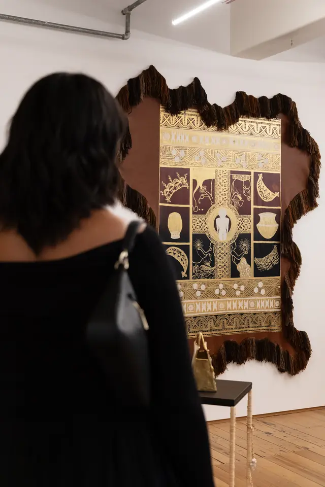 an install shot of showing the back of a figure dressed in black looking at a large artwork on the wall make of textiles and leather. There is a golden handbag sculpture on a plinth in front of the figure.