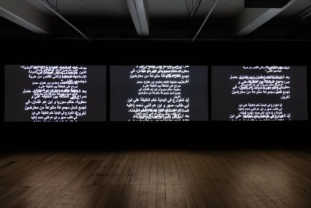 Three projections are illuminated on three projector screens showing scrolling overlayed text in Arabic. The gallery is quite dark and the screens are illuminating the gallery.