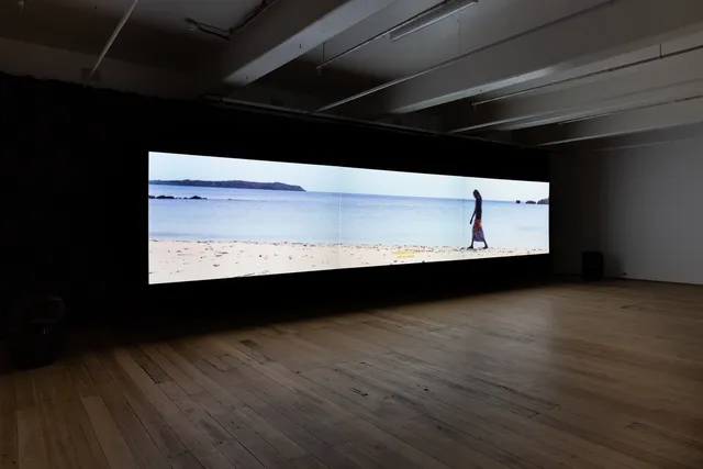 A film is projected across the length of the gallery wall in a long rectangular shape. In the film still, a woman walks across the shoreline of a beach, from right to left.