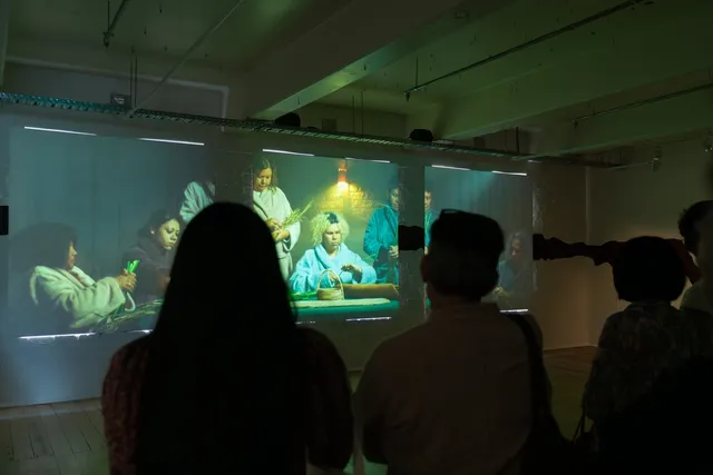 Three people watching video installation, three square projections. Video of people sitting and standing at table weaving in dressing gowns.