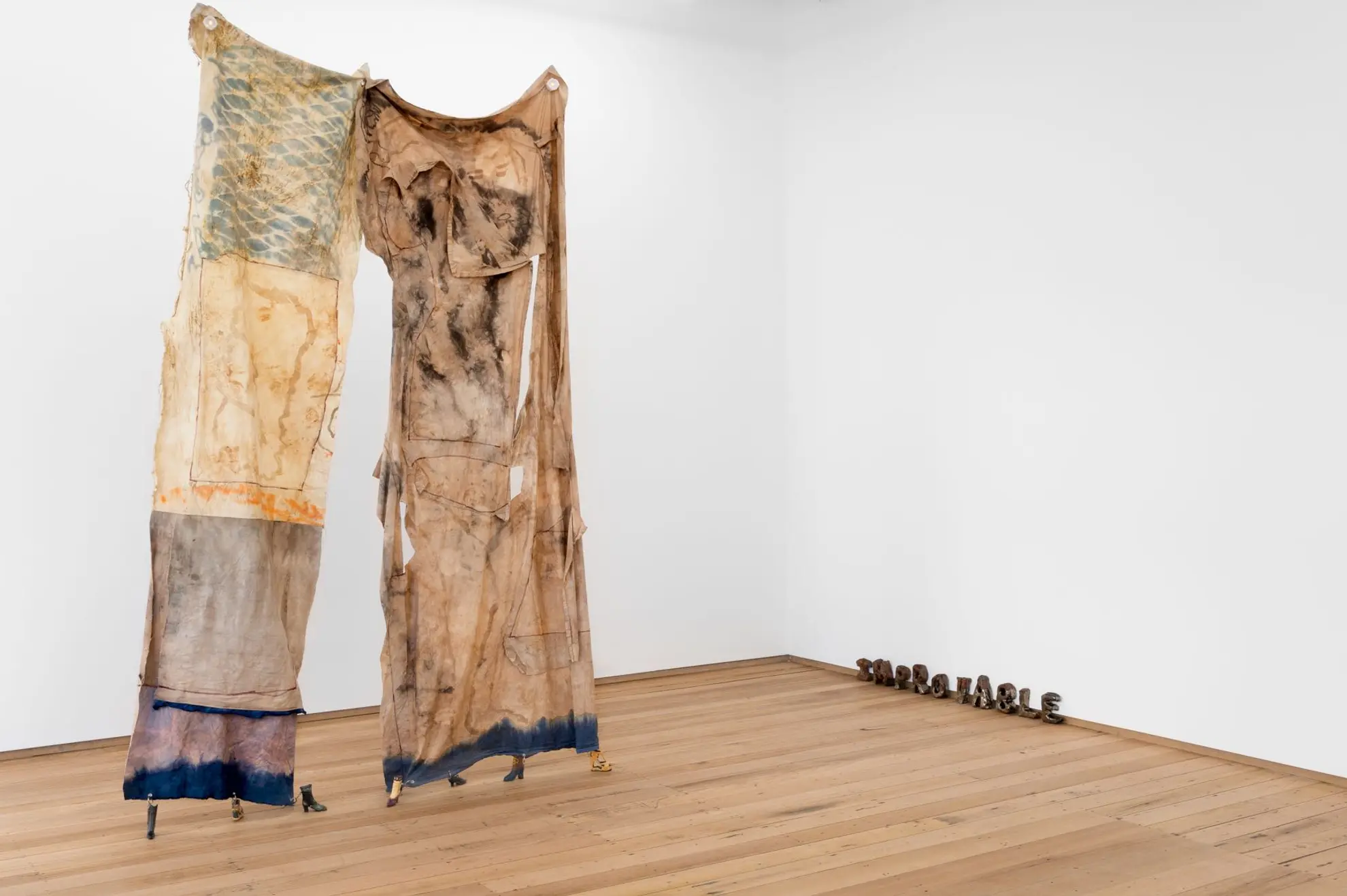 Installation view of Westspace with two large segments of dyed cloth hanging from the ceiling and secured to the ground with a series of small boots. In the bottom right corner sit 10 small handcrafted metal letters that spell out 'Improvable'.