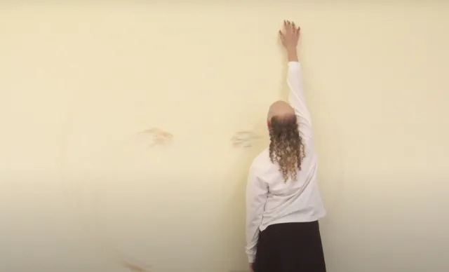 Archie Barry stands In front of a cream coloured wall that has light, gestural pencil marks on it applied by the artist. Archie is reaching up the wall and is wearing a white top and a dark skirt