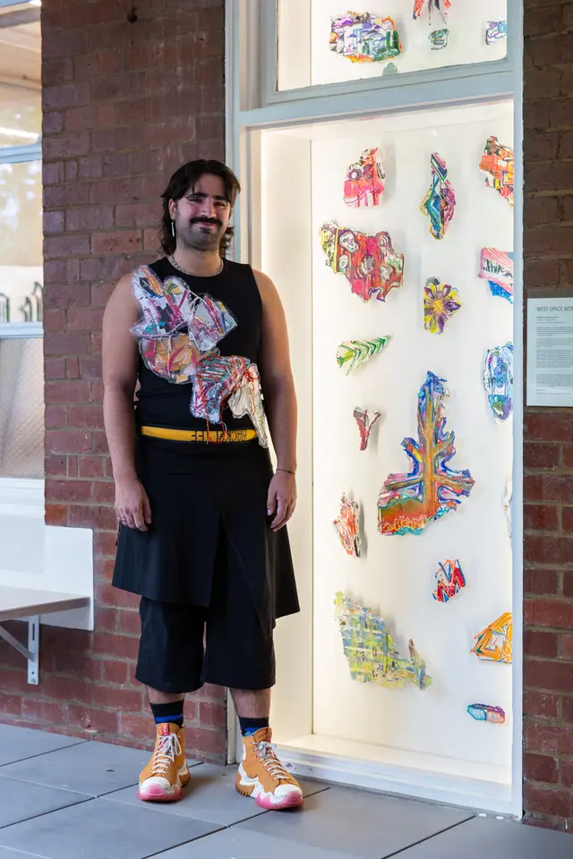 A person stands in front of the west space window. Inside the the window is a collection of collages. The person in front of the window is also wearing these collages on their clothes. They are dressed in black with orange shoes