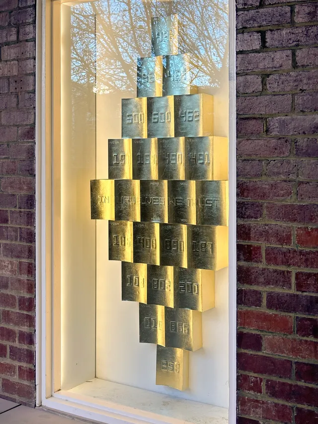 A close up installation view of the west space window. There are a collection of gold rectangular objects that are arranged in a diamond formation. These shapes are emobossed with 3 digit numbers representing the immigration visa codes in Australia
