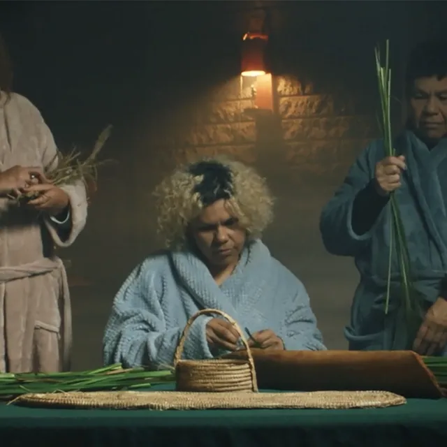 Seven people weaving in a dimly lit room around a green table. Two standing and five sitting with a basket and four candles on the table.
