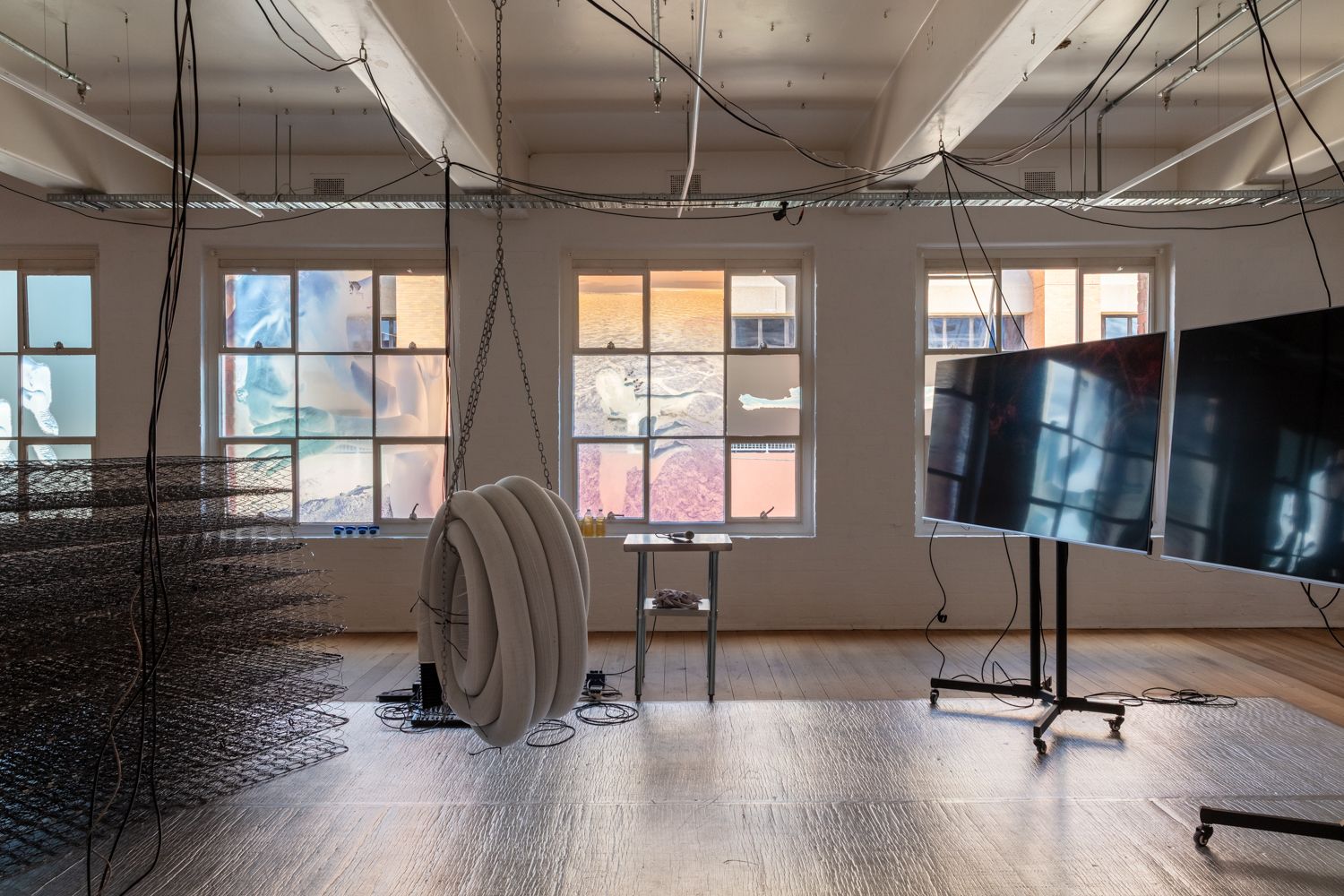 A suspended coil of pipes is hanging from the ceiling,  pile of spring structure of mattresses and two screens are in the gallery. The wooden floor is partially covered with foil mat. The windows are covered with negative photos pf a woman.