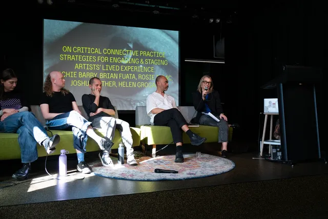 Five individuals sit cross-legged on a green couch on a stage, lit up by a spotlight. A projection screen can be seen behind them.